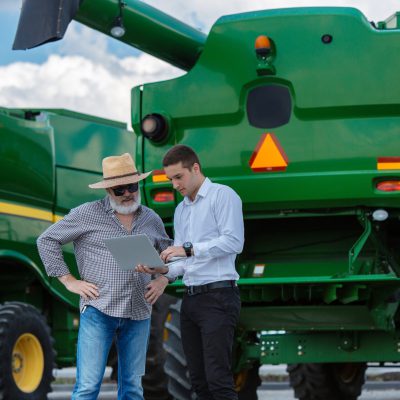 Professional farmer with a modern combine at field in sunlight at work. Confident, bright summer colors. Agriculture, exhibition, machinery, plant production. Senior man near his tractor with investor.